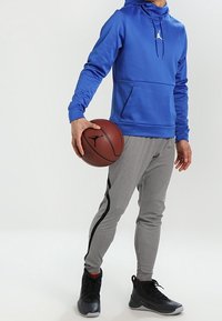 Blauer Kapuzenpullover mit einer vorderen Kängurutasche und einer Kordelzugkapuze, kombiniert mit grauen schmal zulaufenden Jogginghosen mit schwarzen Seitenstreifen und schwarzen Sneakern.