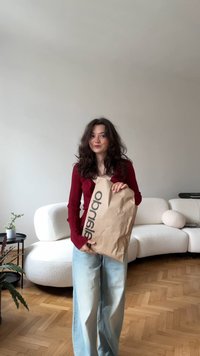 Smeđa papirnata vrećica s logotipom koja se drži u rukama, uz rebrasti crveni pulover i svijetloplave široke traperice, u jednostavnom unutarnjem okruženju.