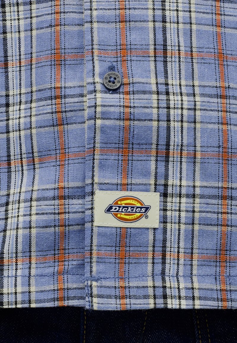 Tessuto a quadri blu, arancione, nero e bianco con un bottone del marchio Dickies e una toppa con logo Dickies ricamata.
