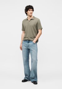 Άνδρας στέκεται με το αριστερό χέρι στην τσέπη, φοράει πουκάμισο πόλο ελιάς, ανοιχτό μπλε παντελόνι με φαρδιά πόδια και μαύρα παπούτσια, κοιτάζοντας προς τα πλάγια.