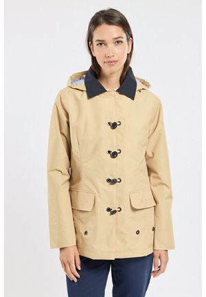 Femme portant un imperméable beige avec des boutons à bascule noirs et un col bleu marine, debout devant un fond blanc uni.