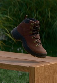 Stivale da trekking in pelle marrone con accenti neri, occhielli dorati e suola robusta, sospeso sopra una superficie di legno con vegetazione sullo sfondo.