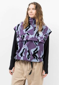 Weste ohne Ärmel aus Fleece in lila und hellblauem Camouflage-Muster, mit hohem Kragen und gesticktem Text, kombiniert mit beigen Hosen.