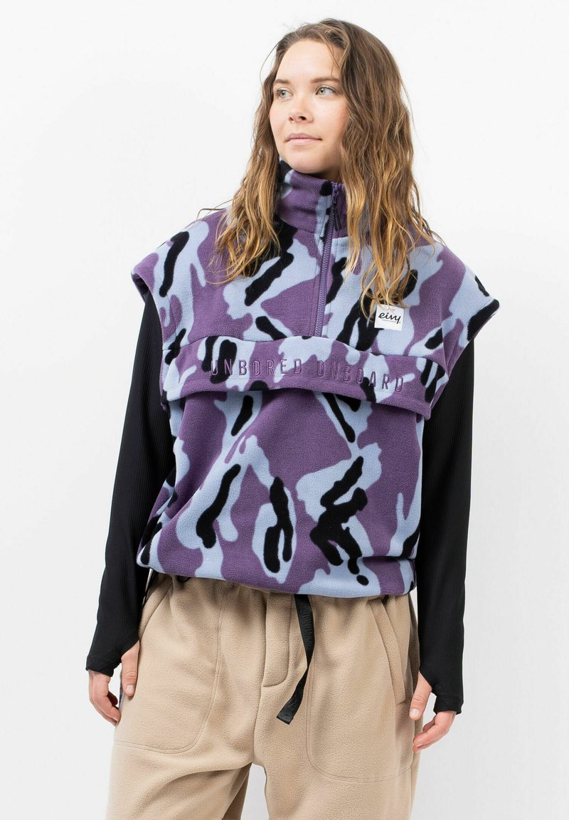 Weste ohne Ärmel aus Fleece in lila und hellblauem Camouflage-Muster, mit hohem Kragen und gesticktem Text, kombiniert mit beigen Hosen.