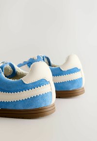 Blauwe suède schoenen met witte zigzag-accenten en een gestructureerde leren hiel, gecombineerd met een bruine rubberen zool. Open achterkant ontwerp.