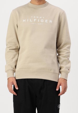 Beigefarbener Tommy Hilfiger Sweatshirt mit weißem Logo-Schriftzug und schwarzen Hosen, getragen von einer Person mit hängenden Armen, schlichter heller Hintergrund.