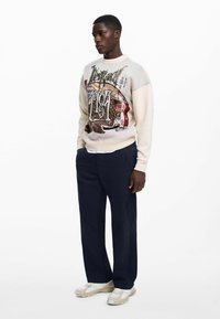 Világos bézs pulóver grafikai mintával és színes kiegészítőkkel, sötét tengerészkék nadrággal és világos sportcipővel párosítva. Simogatós textúra, laza illeszkedés.