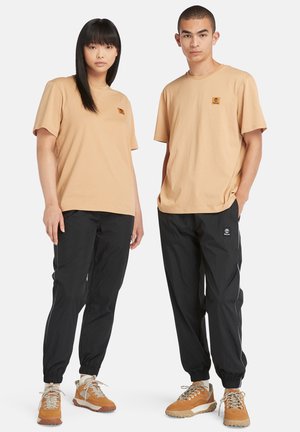 Twee modellen dragen bijpassende beige katoenen T-shirts met kleine logo's en zwarte nylon joggers met zijstreepdelen en elastische manchetten, gecombineerd met beige sneakers.