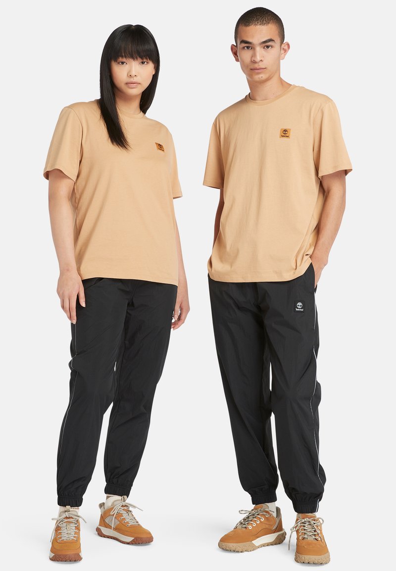 Twee modellen dragen bijpassende beige katoenen T-shirts met kleine logo's en zwarte nylon joggers met zijstreepdelen en elastische manchetten, gecombineerd met beige sneakers.