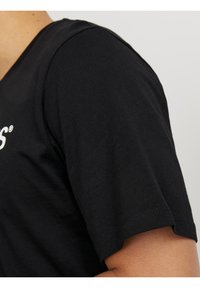 Primer plano de una persona que lleva una camiseta negra de manga corta con texto blanco parcial en el pecho, contra un fondo liso.