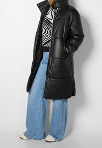 Černá péřová bunda s vysokým límcem, lesklým povrchem a detaily s patentkami, v kombinaci s modrými širokými džíny a světle zbarvenými teniskami.