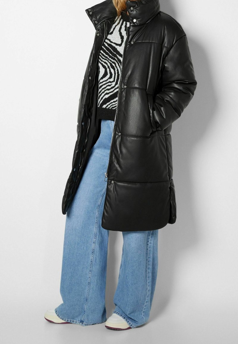 Černá péřová bunda s vysokým límcem, lesklým povrchem a detaily s patentkami, v kombinaci s modrými širokými džíny a světle zbarvenými teniskami.