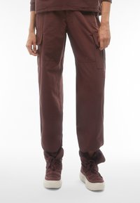 Pantaloni cargo di un rosso scuro, caratterizzati da una vestibilità ampia, tasche laterali e un tessuto testurizzato. Indossati con stivali alla caviglia spessi e allacciati nella stessa tonalità.
