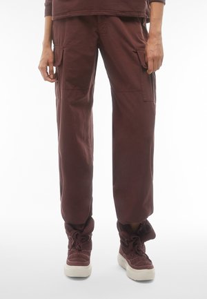 Pantaloni cargo di un rosso scuro, caratterizzati da una vestibilità ampia, tasche laterali e un tessuto testurizzato. Indossati con stivali alla caviglia spessi e allacciati nella stessa tonalità.