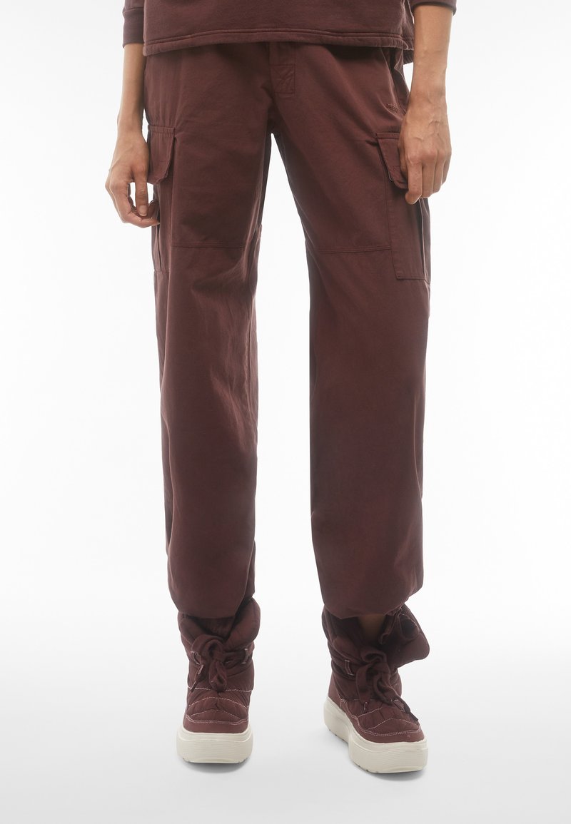 Pantaloni cargo di un rosso scuro, caratterizzati da una vestibilità ampia, tasche laterali e un tessuto testurizzato. Indossati con stivali alla caviglia spessi e allacciati nella stessa tonalità.