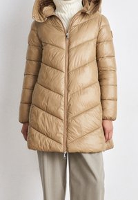 Giacca imbottita beige con un design a chevron, chiusura con zip e cappuccio, abbinata a pantaloni grigi chiari. Tessuto liscio e lucido.
