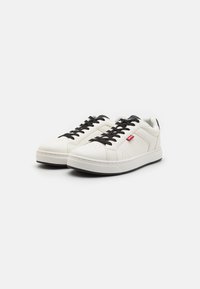 Sneaker bianche in pelle con lacci neri, superficie texturizzata, punta perforata e etichetta logo rossa sul lato. Suola in gomma nera.