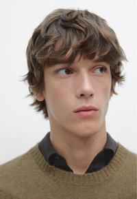 Close-up van een persoon met kort, tousled bruin haar. Draagt een donker button-up shirt onder een olijfgroene gebreide trui.