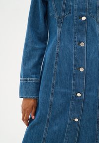 Robe en denim bleu moyen, à manches longues, silhouette droite, dotée de boutons argentés, de coutures contrastées et de deux poches sur la poitrine.