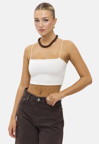 Débardeur blanc court à fines bretelles, porté avec un jean taille haute marron foncé. Présente un collier en perles marron chunky et des boucles d'oreilles en or.