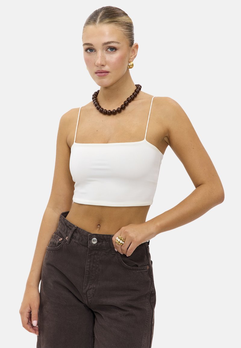 Débardeur blanc court à fines bretelles, porté avec un jean taille haute marron foncé. Présente un collier en perles marron chunky et des boucles d'oreilles en or.