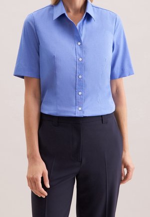 Femme portant une chemise bleu clair à manches courtes boutonnée, rentrée dans un pantalon ajusté bleu marine foncé sur un fond uni.