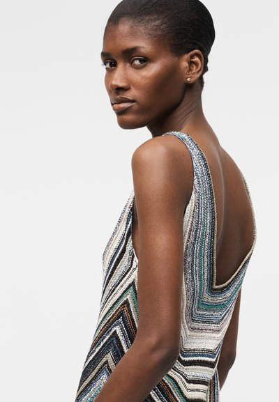 Moteris trumpais plaukais, vilkinti rankovių neturinčią, žemą nugarėlę turinčią su daugiacolorine zigzago juostele suknelę, žiūrinti per petį.