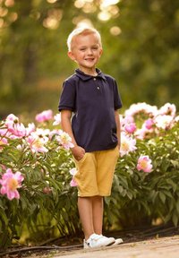 Chłopiec w granatowej koszulce polo z dwoma guzikami, żółtych szortach i białych butach, stojący wśród różowych kwiatów w ogrodzie.