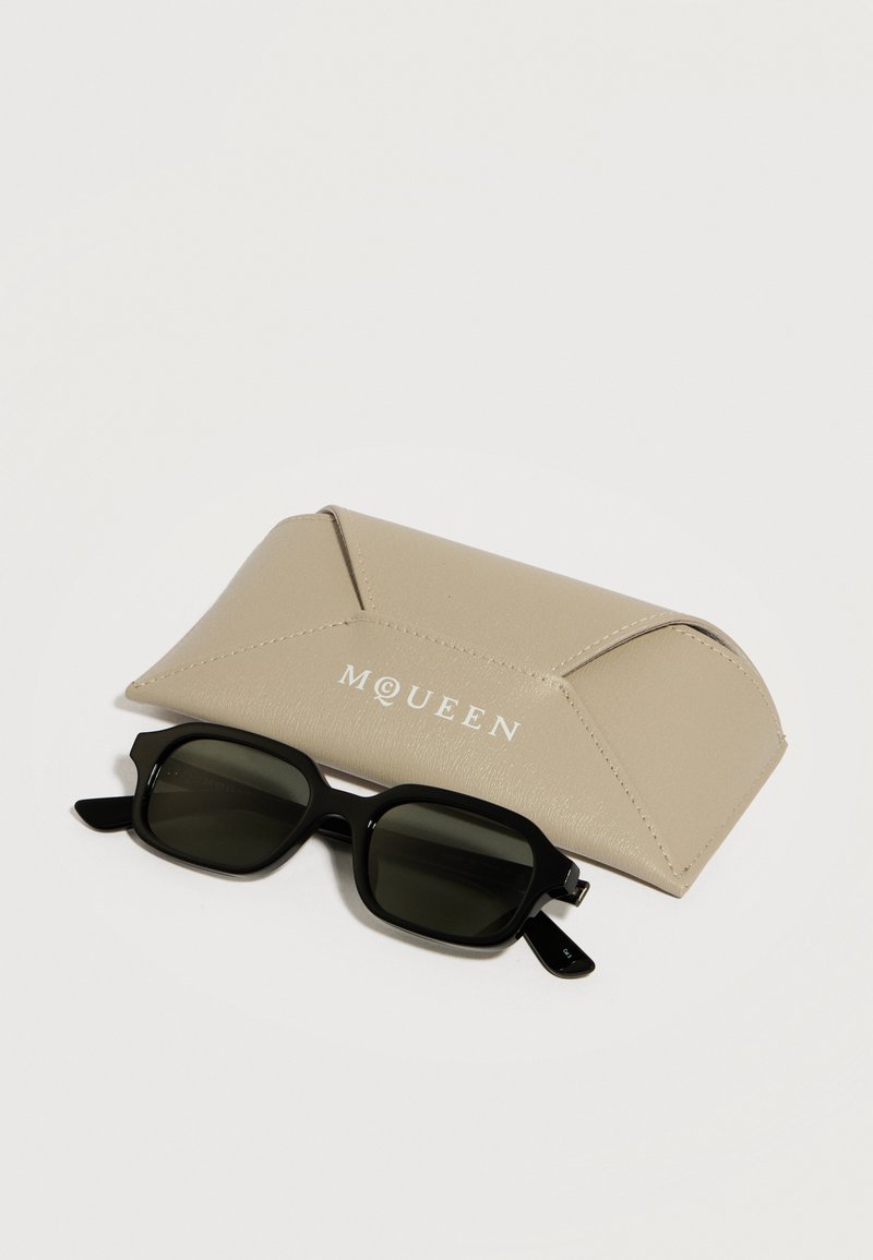 Lunettes de soleil rectangulaires noires avec verres foncés à côté d'un étui à lunettes beige de marque McQueen sur une surface blanche.