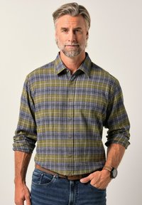 Středně starý muž se šedými vlasy a vousy, oblečený v zelenošedé kostkované košili s knoflíky a modrých džínách, stojící s jednou rukou v kapse.