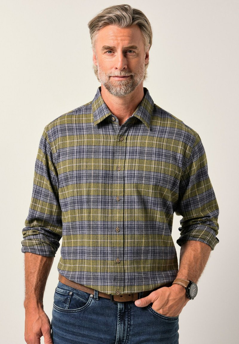 Středně starý muž se šedými vlasy a vousy, oblečený v zelenošedé kostkované košili s knoflíky a modrých džínách, stojící s jednou rukou v kapse.