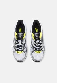 Scarpe da ginnastica bianche con lacci neri, dettagli gialli e una parte superiore in mesh. Presentano un colletto imbottito e una suola testurizzata.