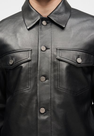 Μαύρο δερμάτινο μπουφάν με ασημένια κουμπιά τύπου snap, μυτερό γιακά και δύο θήκες στο στήθος με κουμπιά.