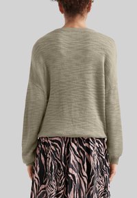 Olivgrüner, gestrickter Pullover mit lockerer Passform und überschnittenen Schultern, kombiniert mit einem plissierten Rock in Schwarz und Pink mit Zebra-Streifen.