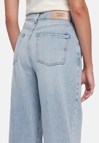 Jeans en denim bleu clair à jambes larges avec poches arrière, taille haute et une étiquette en cuir. Texture douce avec une apparence délavée.