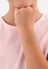 Bracciale in argento con un accento a forma di cuore e decorazioni rosa, indossato su un polso contro uno sfondo di camicia rosa chiaro.