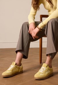 Gele suède sneakers met witte veters, ronde neus en beige rubberen zool, met een gemusterde textuur en een zijlogo-detail.