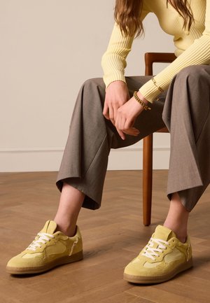 Gele suède sneakers met witte veters, ronde neus en beige rubberen zool, met een gemusterde textuur en een zijlogo-detail.