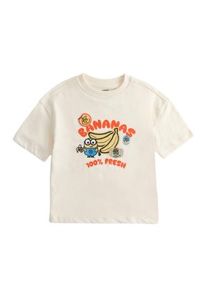 Barn iført en cremefarvet T-shirt med tegneserie bananer og tekst, denimshorts, stribede sokker og farverige sneakers står på en ensfarvet baggrund.