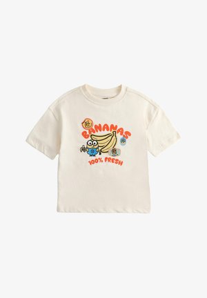 Małe dziecko ubrane w kremowy t-shirt z rysunkowymi bananami i napisem, dżinsowe szorty, paskowane skarpetki i kolorowe tenisówki, stojące na jednolitym tle.