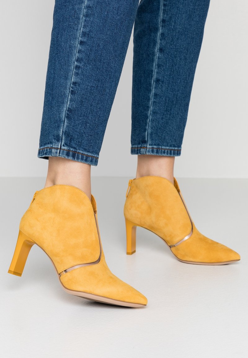 Femme portant un jean denim bleu moyen et des bottines à talons hauts en daim jaune, debout sur un sol gris clair.