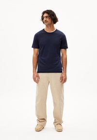 T-shirt navy a maniche corte, collo a giro, abbinato a pantaloni beige. Il tessuto sembra liscio e leggero. Il modello sta in piedi con le mani ai fianchi su uno sfondo bianco.