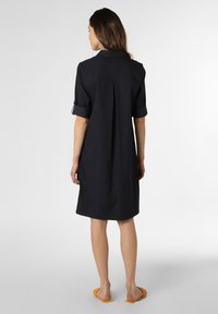 Marineblaues Hemdkleid mit kurzen umgeschlagenen Ärmeln, gefälteter Rückseite und lockerem Schnitt, aus einem glatten Stoff gefertigt. Dazu werden gelbe flache Sandalen getragen.