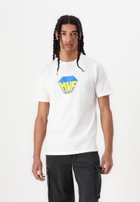 Hvid bomulds T-shirt med et blåt og gult sekskantet logo med "HUF" tekst. Modellen bærer den med sorte cargo shorts, hænderne langs siderne.