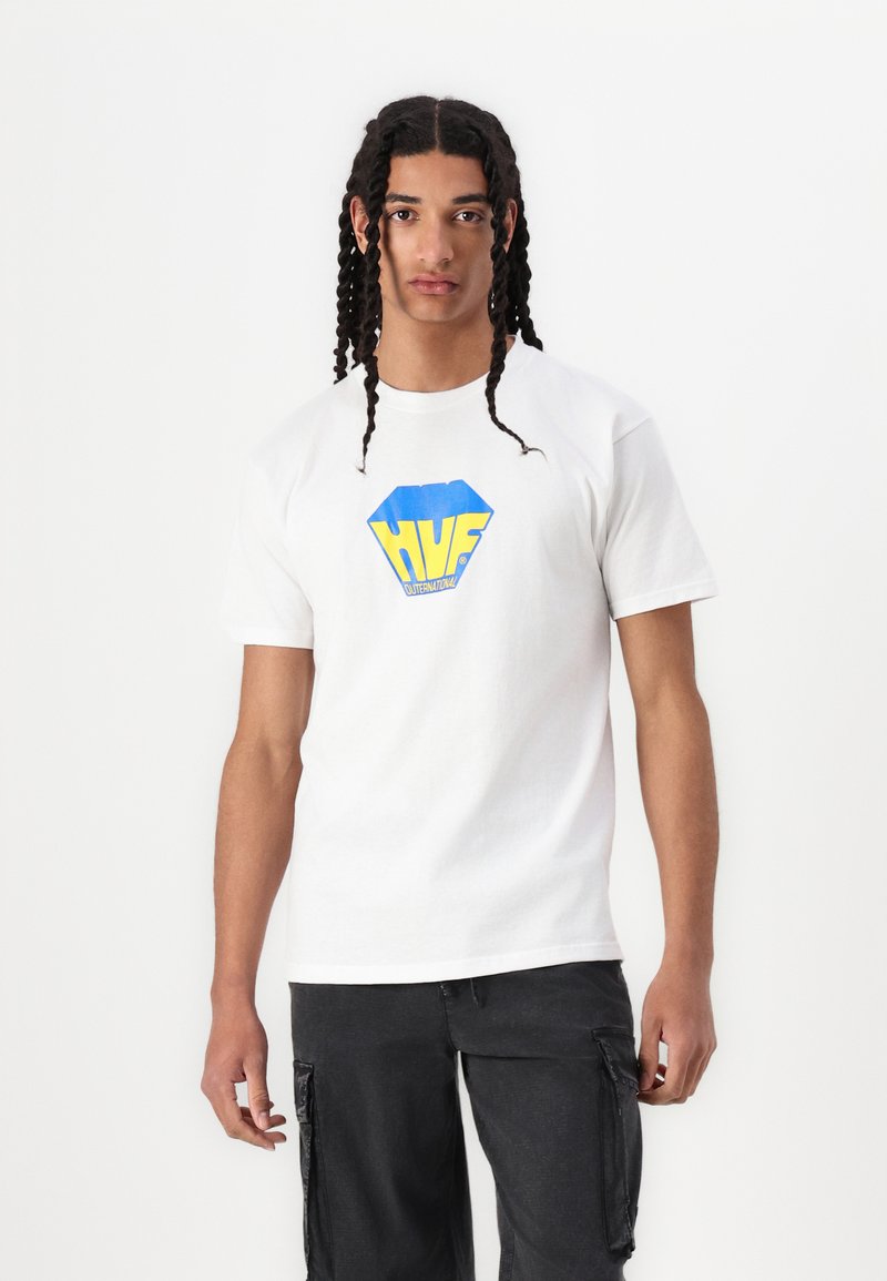 Hvid bomulds T-shirt med et blåt og gult sekskantet logo med "HUF" tekst. Modellen bærer den med sorte cargo shorts, hænderne langs siderne.