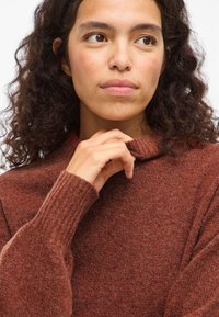 Göndör, sötét hajú nő, aki rozsdaszínű kötött pulóvert visel, óvatosan fogja a gallért, miközben kissé oldalra néz.