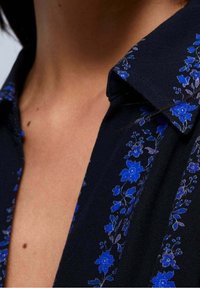 Schwarzes Hemd mit blauer Blumenstickerei am Kragen und an den Schultern; der Stoff wirkt weich und hat eine glatte Textur.