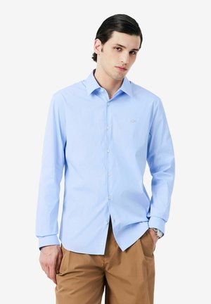 Junger Mann trägt ein hellblaues Hemd mit Knöpfen und hellbraune Hose, mit einer Hand in der Tasche und neutralem Gesichtsausdruck.