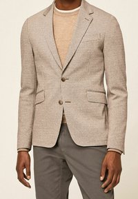 Beige Hahnentritt-Blazer mit Reverskragen, zwei Knöpfen und Seitentaschen; darüber ein hellbeiger Pullover getragen und mit grauer Hose kombiniert.