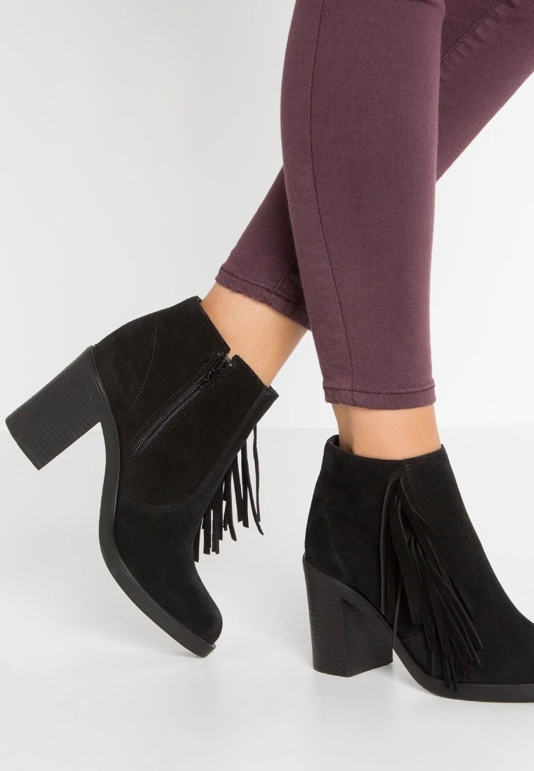 Femme portant des bottines en daim noir avec talons carrés et fermetures éclair latérales, ornées de franges décoratives, associées à un pantalon violet raccourci.